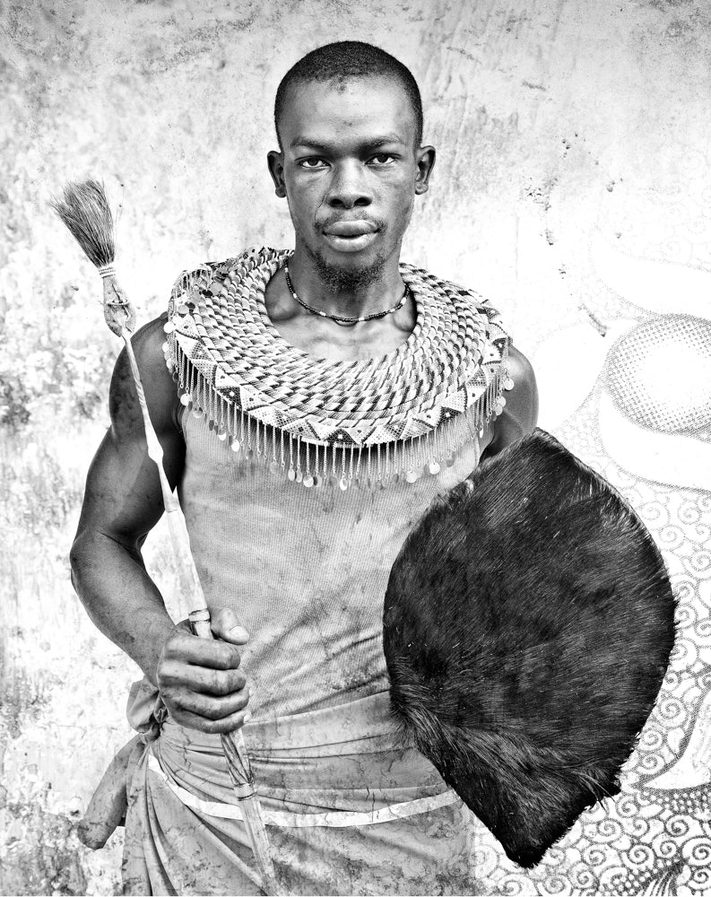 Black and white photograph of a Masai warrior holding a spear and wearing a traditional headdress and shield.